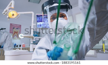 Dentist in coverall leaning over patient putting oxigen mask before surgery in stomatological office with new normal. Stomatolog wearing safety gear against covid19 during heatlhcare check of person. Royalty-Free Stock Photo #1883195161