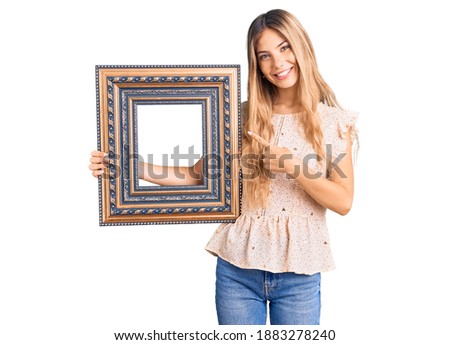 Beautiful caucasian woman with blonde hair holding empty frame smiling happy pointing with hand and finger 