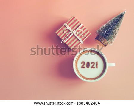 Cup of coffee with the number 2021 on frothy surface over pink background with gift box and Christmas tree. Holidays food creative concept for active days in New Year 2021. (top view, space for text)