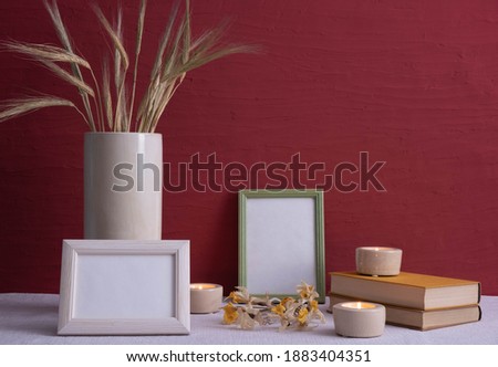 old books, candles are burning, a gray vase with ears of corn, dry daffodils, photo frames on a red background on the table
