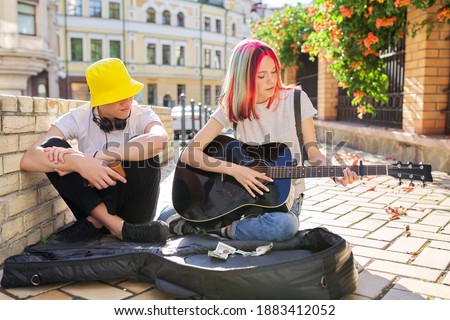 Couple of teenage hipsters street musicians making money by music, girl with colored hair singing and playing acoustic guitar. Teenagers sitting on sidewalk on city street