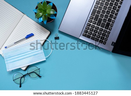 On the office table, a laptop, glasses, a pen, a notebook and a medical mask to protect against viruses on a blue background. Remote work concept during a pandemic. Flat lay. copy space. top view