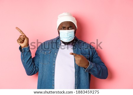 Coronavirus, lifestyle and global pandemic concept. Angry and disappointed african-american man in face mask pointing left, staring at camera displeased, pink background