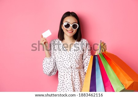 Beautiful asian woman in sunglasses going shopping, holding bags and showing credit card, standing over pink background