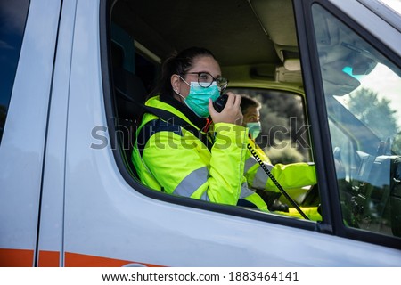 Portrait of a young woman wearing a mask and uniform in an ambulance during a volunteer shift answering an emergency call with her fellow driver - Concept of pandemic from Covid-19, Coronavirus Royalty-Free Stock Photo #1883464141