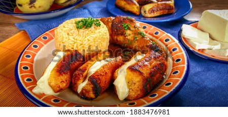 Delicious Ecuadorian dish of fried plantains with melted cheese inside, accompanied by brown rice and chicken, all on a wooden table and a blue table cloth.  Royalty-Free Stock Photo #1883476981