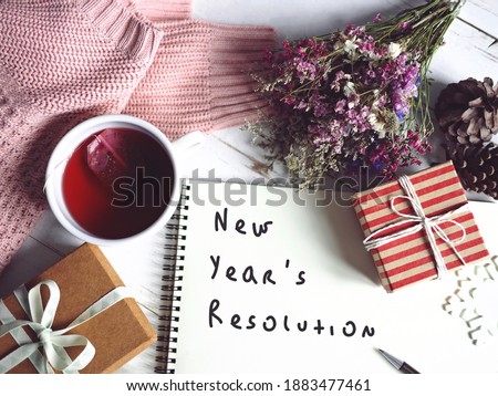 New Year's resolution on notebook paper with pen, gift box, snowflake over white painted wood planks background with cup of tea, pink knit sweater, flower bunch, pine cones. Goal setting theme.