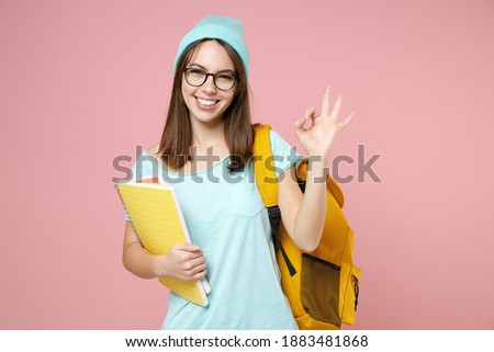 Funny young woman student in blue t-shirt hat glasses backpack hold notebooks showing ok okay gesture isolated on pink background studio portrait. Education in high school university college concept