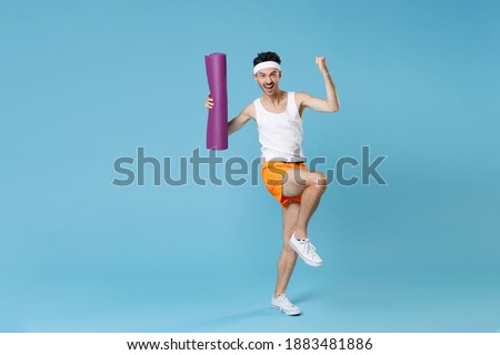 Full length portrait joyful young sporty man with skinny body sportsman in headband shirt shorts hold yoga mat doing winner gesture isolated on blue background. Workout gym sport motivation concept