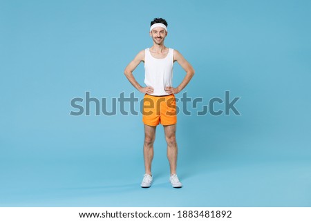 Full length portrait smiling young fitness man with skinny body sportsman in headband shirt shorts standing with arms akimbo on waist isolated on blue background. Workout gym sport motivation concept Royalty-Free Stock Photo #1883481892