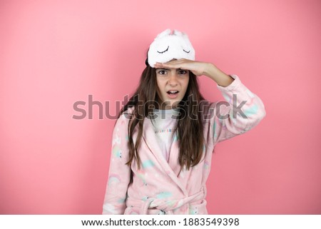Pretty girl wearing pajamas and sleep mask over pink background looking far away with hand over head. Searching concept.