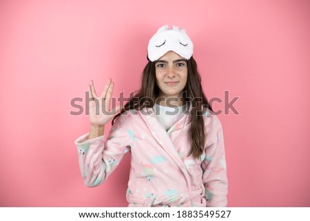 Pretty girl wearing pajamas and sleep mask over pink background doing hand symbol