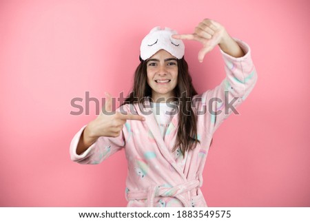 Pretty girl wearing pajamas and sleep mask over pink background smiling making frame with hands and fingers with happy face