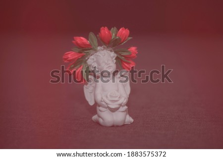 Figurine of a white angel with a rose on a gray background. On the head there is an ornament in the form of a wreath of red flowers.
