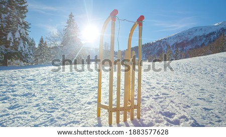 LENS FLARE: Wooden sled is illuminated by the golden winter sun rays while standing upright in the middle of a snowy meadow in Kranjska Gora. Sleigh is set aside during a fun day in wintry mountains