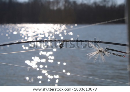 A rooster tail fishing lure sits ready on a fishing pole with a lake and wintery forest in the background.