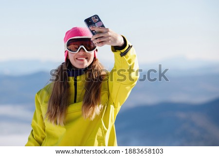 Woman in ski clothes takes a selfie against the backdrop of blue mountains. Yellow or mustard jacket with a hood, a knitted hat, ski goggles. Healthy lifestyle. Sports concept. Selective focus.