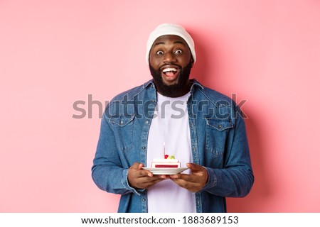 Cheerful african-american guy celebrating birthday, making wish on bday cake with lit candle, smiling happy, standing over pink background