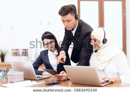 A man in headphones explains something to workers at a computer. Women in headphones work in the office with laptops. 