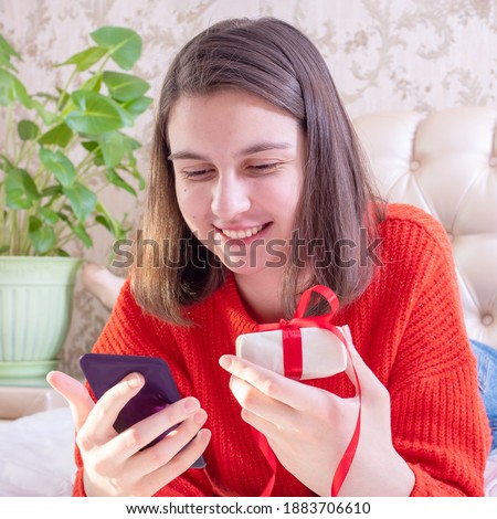 Happy young girl in a red sweater holding a gift, congratulating family or friends with a smartphone, close-up, front view, square. Online greeting concept. Stay at home concept