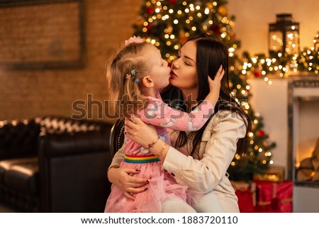 Pretty young mother woman with little daughter girl in fashionable clothes kiss on the background of christmas decorations and lights. Family winter holidays 