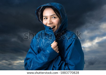 Portrait of a smiling girl dressed in blue raincoat in drops posing with hood against the background of the dramatic sky. Bad weather concept.