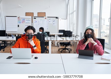 Smart man wearing protective mask studying at designer faculty sitting at the desk near his group mate, opposite of his laptop. Man and woman keeping a social distancing. Stock photo