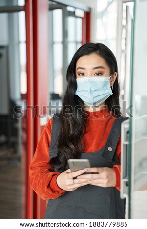 Successful female student with dark hair wearing protective mask sharing her success with his friends via social networks after passed exam while standing at comfortable college hall. Stock photo