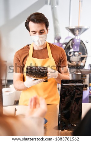 Portrait of the seller wearing apron holding an organic coffee bag after the filling it from a bulk dispenser. Man working at the coffee store. Stock photo