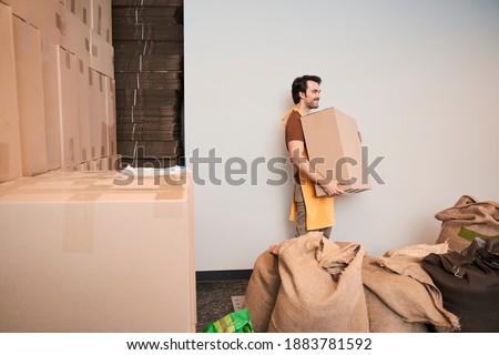 Side view cheerful unshaven professional coffee roaster worker holding cupboard box in hands while going at factory with cup board boxes at the background. Industry concept