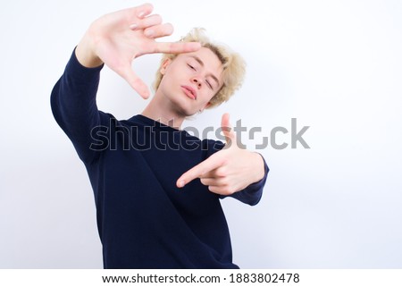 Young handsome Caucasian blond man standing against white background making finger frame with hands. Creativity and photography concept.