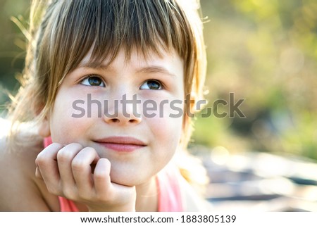 Portrait of pretty child girl with gray eyes and long fair hair leaning on her hands smiling happily outdoors on blurred bright background. Cute female kid on warm summer day outside.