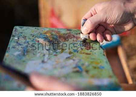 Close-up picture of young male hand, squeezing oil paint from small tube on artist paint palette. Painting workshop in rural countryside. Artistic education modern concept. Outdoors leisure activities