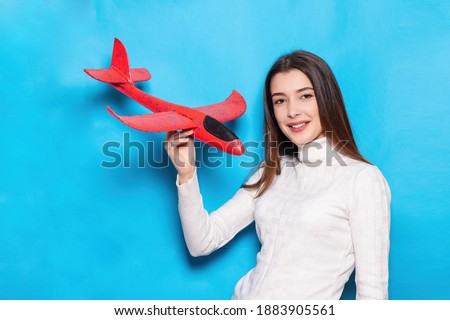 Cute young woman with a smile in striped sweater. holds plane in her hands and points a finger at it on isolated blue background. Emotional face. Travel concept, air flight, airplane tickets