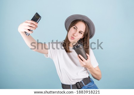 Smiling adorable caucasian female making selfie photo on smartphone with positive expression in casual clothing and coffee in gands over blue background.