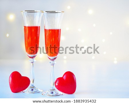 Two beautiful glasses with delicate engraving and red hearts are filled with red drink and stand on a blurred background with a side of the garland, a horizontal image with a soft focus and a place f