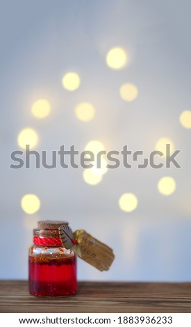 A small glass jar with red-gold liquid and a gold tag stands on a wooden table on a blur of a table with a side from a garland for valentines day, a vertical image with a soft focus, copyspace