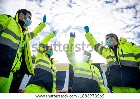 Portrait of a group of paramedics at the end of their shift in front of the ambulance while they high-fives in the air dressed in uniform and wearing a mask for protection from Coronavirus, Covid-19 Royalty-Free Stock Photo #1883973565