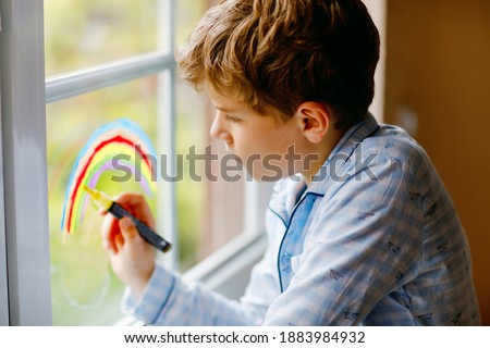 Lovely school kid boy in pajamas painting rainbow with colorful window color during pandemic coronavirus quarantine. Children painting rainbows around the world with the words Let's all be well.