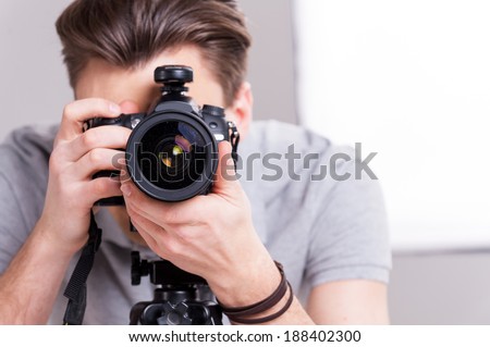 Smile! Young man focusing at you with digital camera while standing in studio with lighting equipment on background  Royalty-Free Stock Photo #188402300