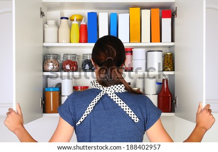 A woman seen from behind opening the doors to a fully stocked pantry. The cupboard is filled with various food stuff and groceries all with blank labels. Horizontal format the woman is unrecognizable. Royalty-Free Stock Photo #188402957