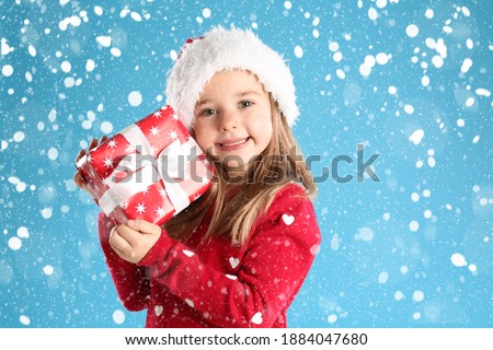 Cute child in Santa hat with Christmas gift under snowfall on light blue background