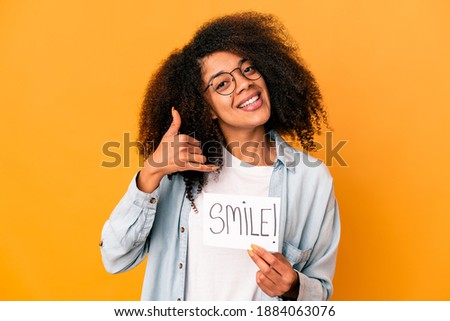 Young african american curly woman holding a smile message placard showing a mobile phone call gesture with fingers.