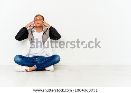 Young caucasian man sitting on the floor focused on a task, keeping forefingers pointing head.