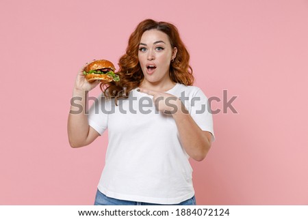 Shocked young redhead plus size body positive woman 20s in white casual t-shirt pointing index finger on american classic fast food burger isolated on pastel pink color background studio portrait