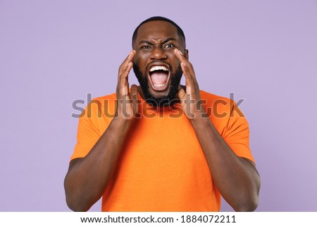 Crazy young african american man 20s wearing basic casual orange blank empty t-shirt screaming with hands gesture near mouth looking camera isolated on pastel violet colour background studio portrait