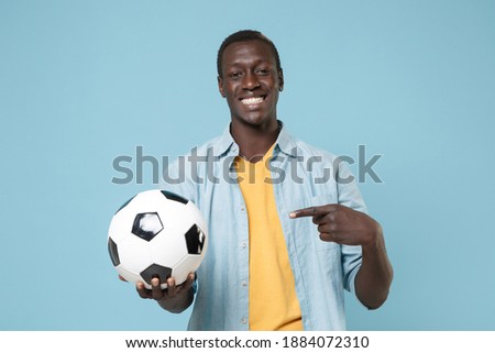 Smiling young african american man football fan player in casual shirt yellow t-shirt isolated on blue background. Sport leisure lifestyle concept. Playing football point index finger on soccer ball