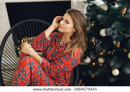 Side view of girl wears festive pajamas and having fun at home near Christmas Tree. Smiling woman sit on black chair in red night-wear