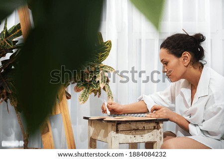 A beautiful concentrated woman artist squeezing the oil paint from the tube into the palette, sitting in a front of easel with canvas.