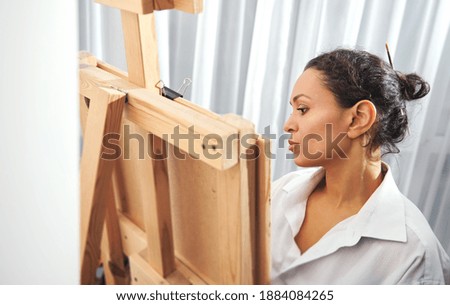 Side view of a beautiful brunette painter with gathered hair in a bun and paint brushes in her hair standing in front of the easel and drawing